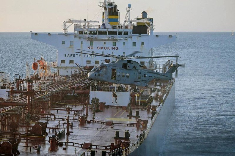 A helicopter hovers above the deck of a large cargo ship at sea, with crew members standing on the deck below and "NO SMOKING" and "SAFETY FIRST" signs visible on the ship's superstructure.