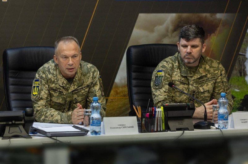 Two men in Ukrainian military camouflage uniforms sit at a conference table with microphones, water bottles, and nameplates, one speaking while the other listens with eyes closed.