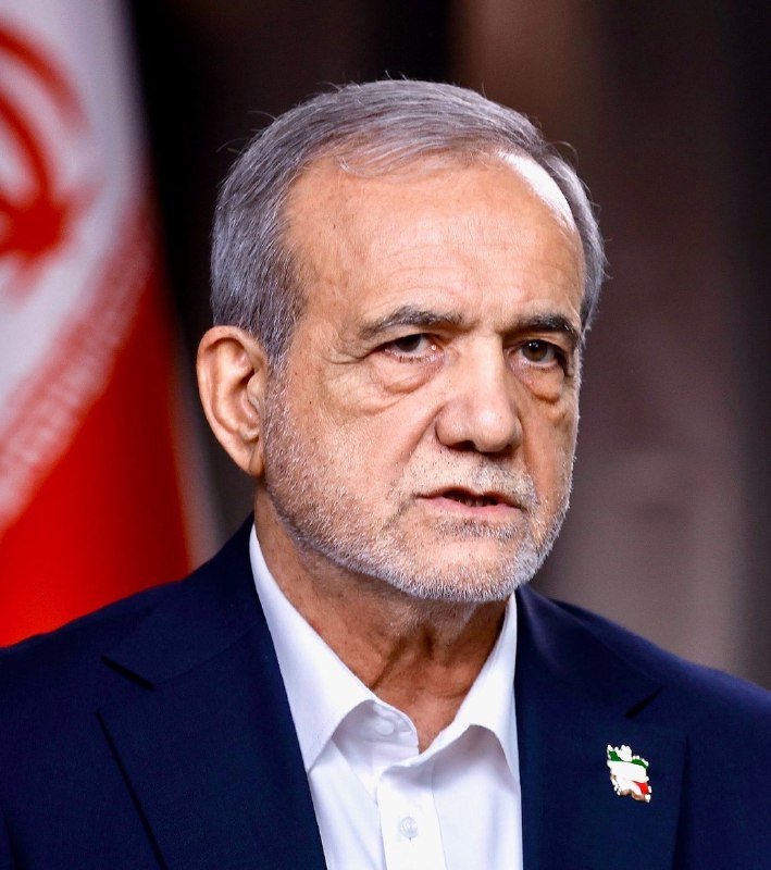 A gray-bearded man in a dark blue suit and white shirt stands before a red and white flag, with a small map pin on his lapel.