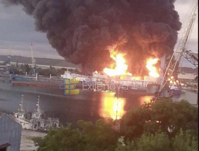 Large flames and thick black smoke rise from a docked ship in a harbor, reflecting intensely on the water below.