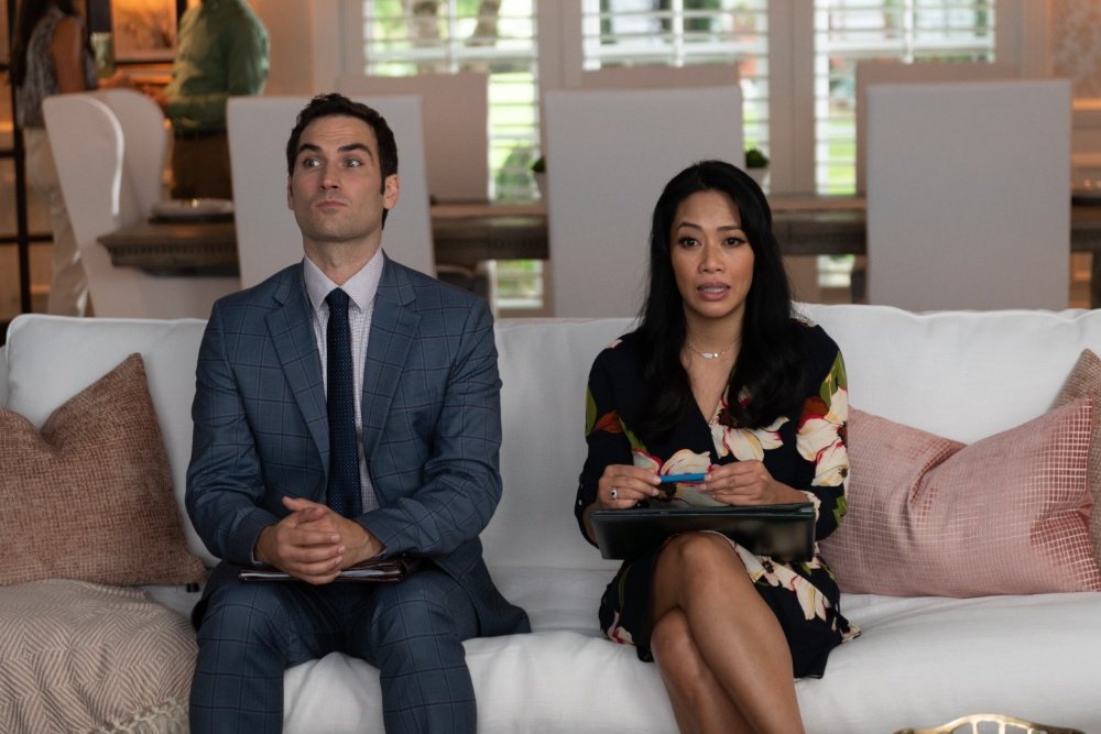 This appears to be a scene from a television show or film featuring a man in a suit and a woman in a floral dress seated on a couch in a living room setting.