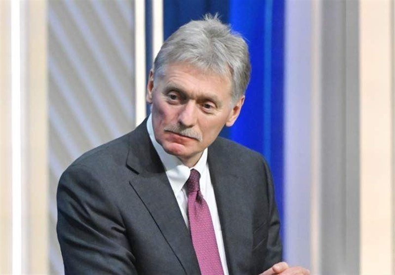 A gray-haired man with a mustache wearing a dark suit, white shirt, and magenta tie sits against a blue and beige backdrop.