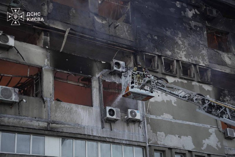 A firefighter in a basket of an extended ladder truck sprays water onto a smoke-damaged multi-story building with shattered windows and a logo reading "ДСНС КИЄВА."