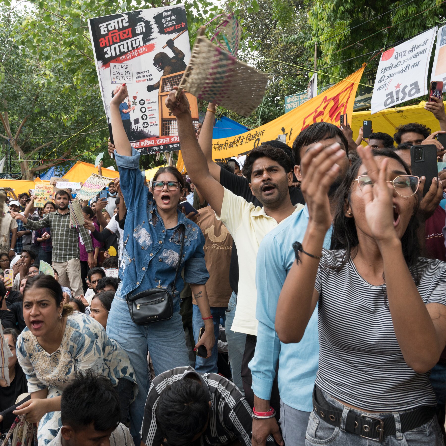 A large crowd of demonstrators chants and holds up Hindi-language protest signs and placards during an outdoor rally beneath trees.