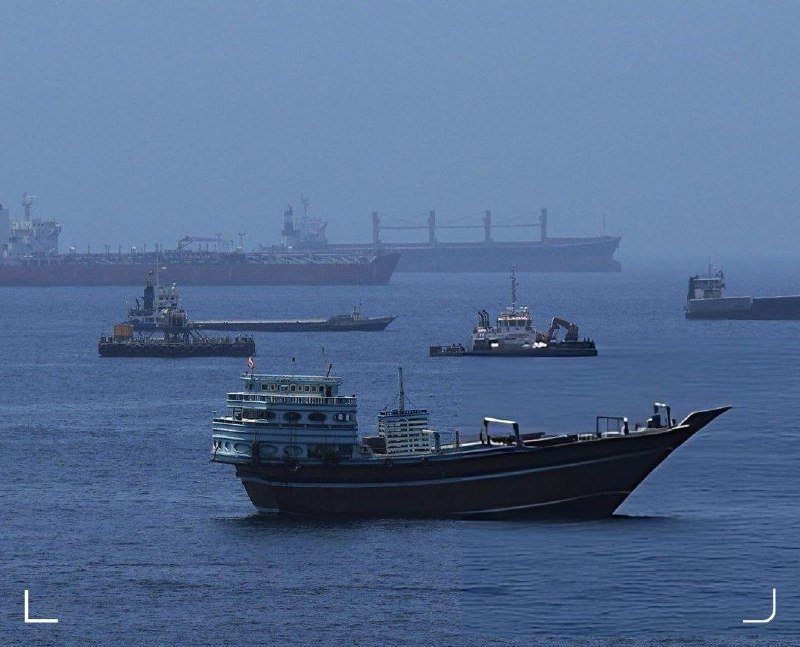 A wooden cargo vessel sails on calm blue waters, with two smaller tugboats and two large ships visible in the hazy background.