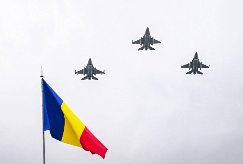 Three military fighter jets fly in formation across an overcast sky above a blue, yellow, and red flag.