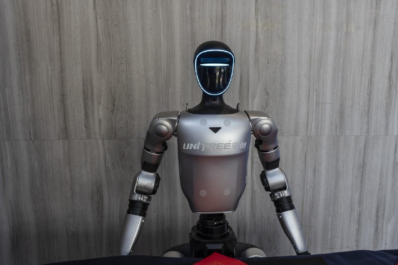 A silver humanoid robot with a dark visor and the text "UNITREE" on its chest stands against a gray wood-paneled wall.