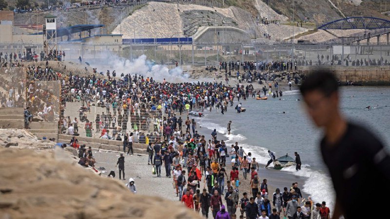 A large crowd gathers on a beach and along a fenced border area as white smoke rises in the background.