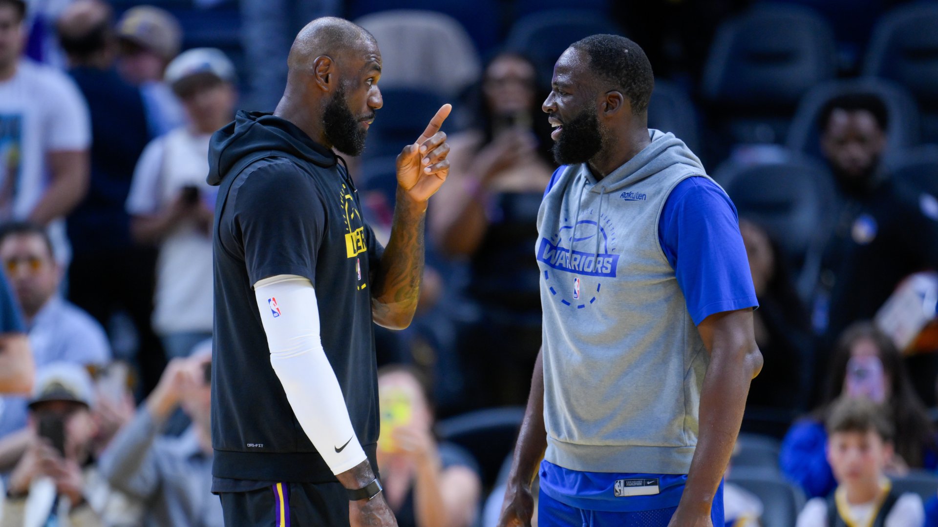 Two basketball players, one in a black Lakers hoodie and the other in a gray Warriors shirt, share a laugh during pregame warmups on the court.