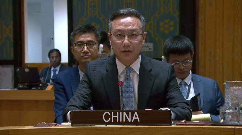 A man in a dark suit and glasses speaks at a desk bearing a "CHINA" nameplate, with other formally dressed individuals seated behind him in a conference room.