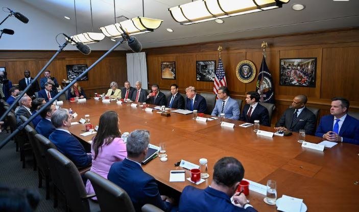 A group of people in business attire sit around a large wooden conference table in a wood-paneled room featuring American flags and a presidential seal on the wall.