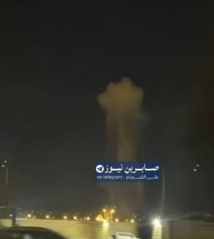 A large plume of smoke rises into a dark night sky above a lit roadway, with vehicles visible in the foreground.