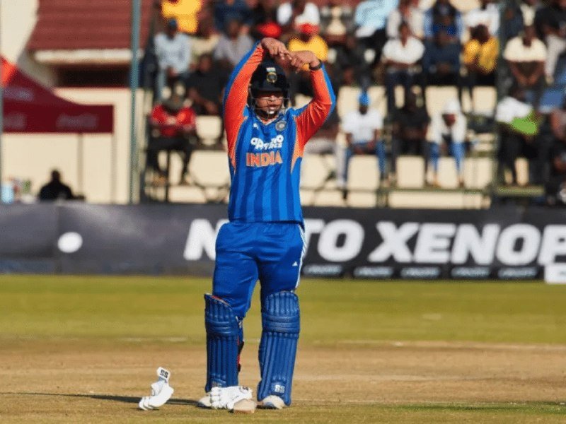 A cricket player wearing a blue and orange "INDIA" jersey, helmet, and pads stands on the pitch with both arms raised, while spectators watch from the stands.