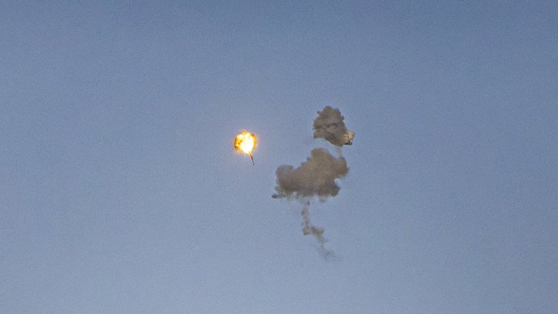 A bright orange fireball streaks through a clear blue sky, leaving a dark gray smoke plume trailing behind it.