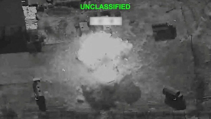 A grayscale aerial surveillance image marked "UNCLASSIFIED" in green shows a large white smoke plume rising from a built-up area with scattered structures visible below.