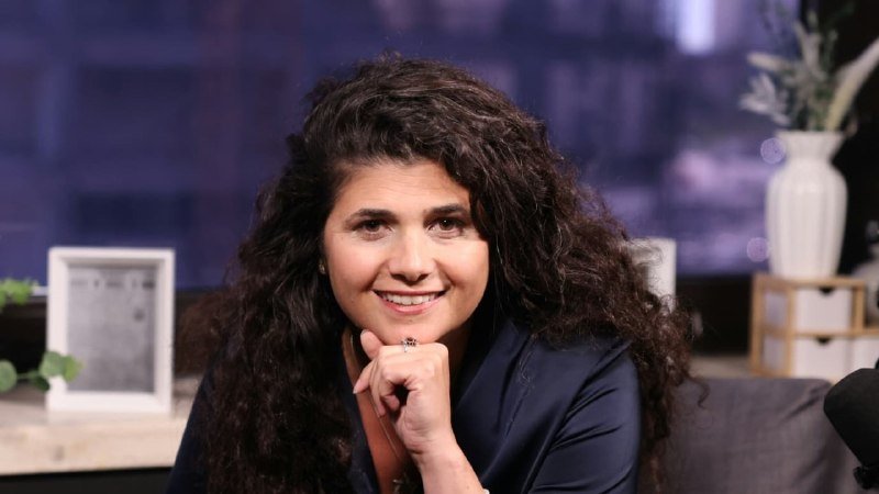 A smiling woman with long dark curly hair sits resting her chin on her hand, wearing a dark blazer in a studio setting with a blurred background.