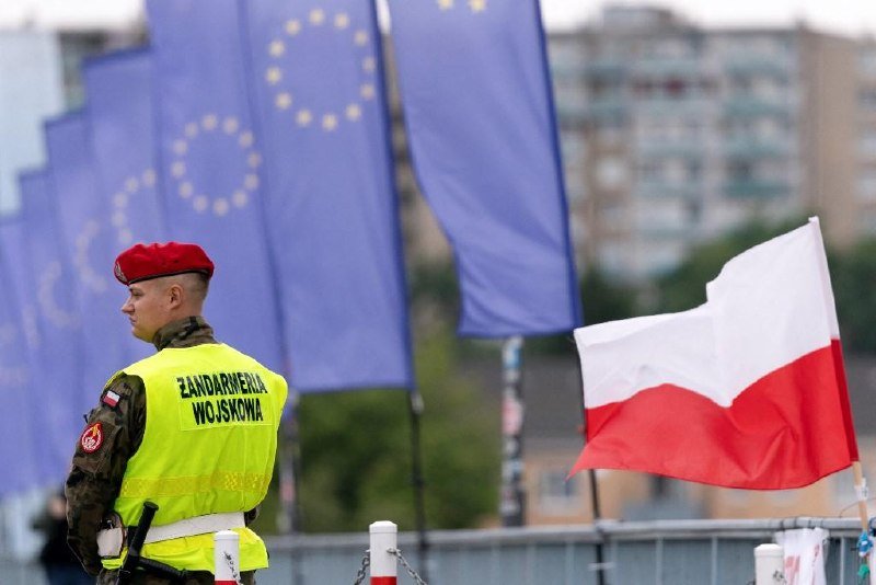 A military police officer wearing a "Żandarmeria Wojskowa" vest and red beret stands near a barrier, with EU and Polish flags visible in the background.