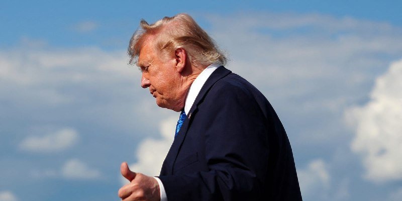 A man with blonde hair wearing a dark suit and blue tie walks outdoors against a partly cloudy sky, gesturing with his hand.
