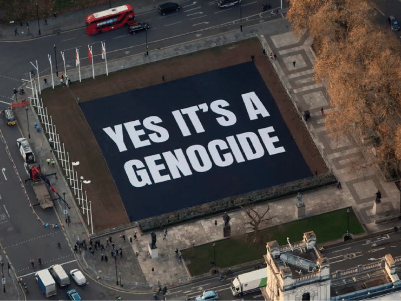 A large black banner displaying the white text "YES IT'S A GENOCIDE" is laid out on a paved public square, surrounded by streets, vehicles, and pedestrians.