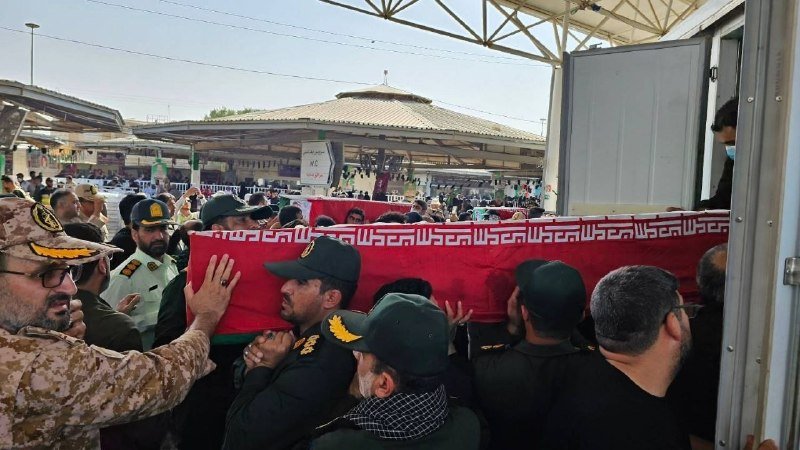 Uniformed men in military and dark clothing carry a red-draped coffin through a crowded outdoor area toward an open doorway.