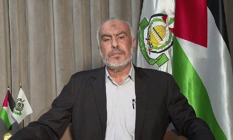 A bearded man in a dark suit and light shirt sits before draped curtains, flanked by two flags bearing a green, white, and black emblem.