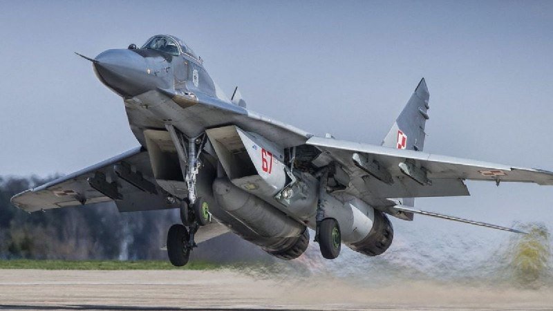 A gray military fighter jet with red and white tail markings lifts off a runway, landing gear extended, against a clear sky with trees in the background.
