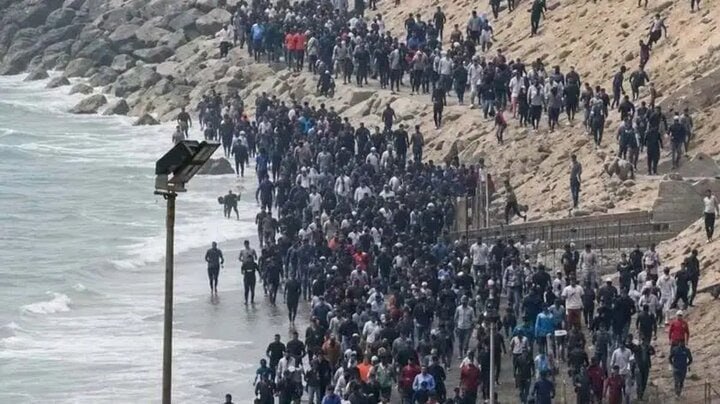 A large crowd of people walks along a narrow coastal strip beside the ocean, with waves on one side and a sandy embankment on the other.