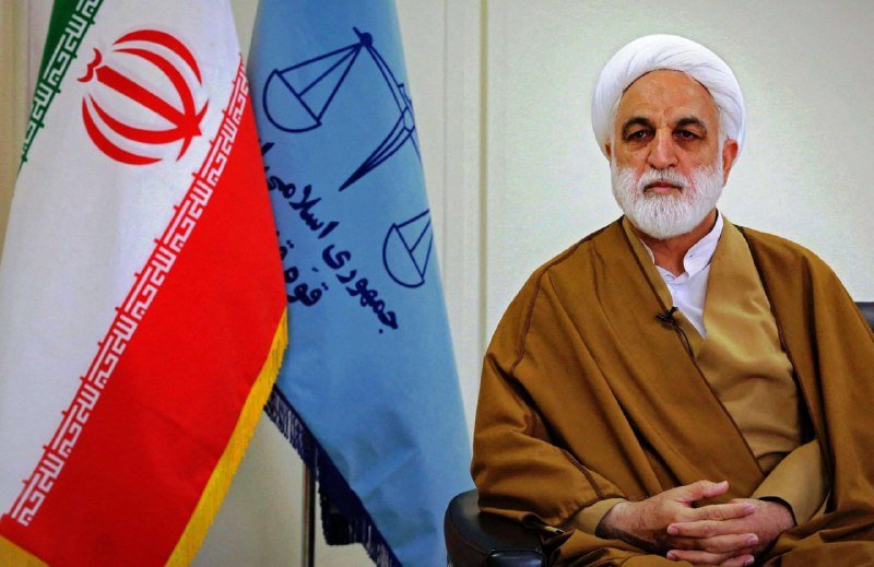 A bearded man in a white turban and brown clerical robe sits beside the Iranian flag and a blue emblem featuring scales of justice and Persian script.