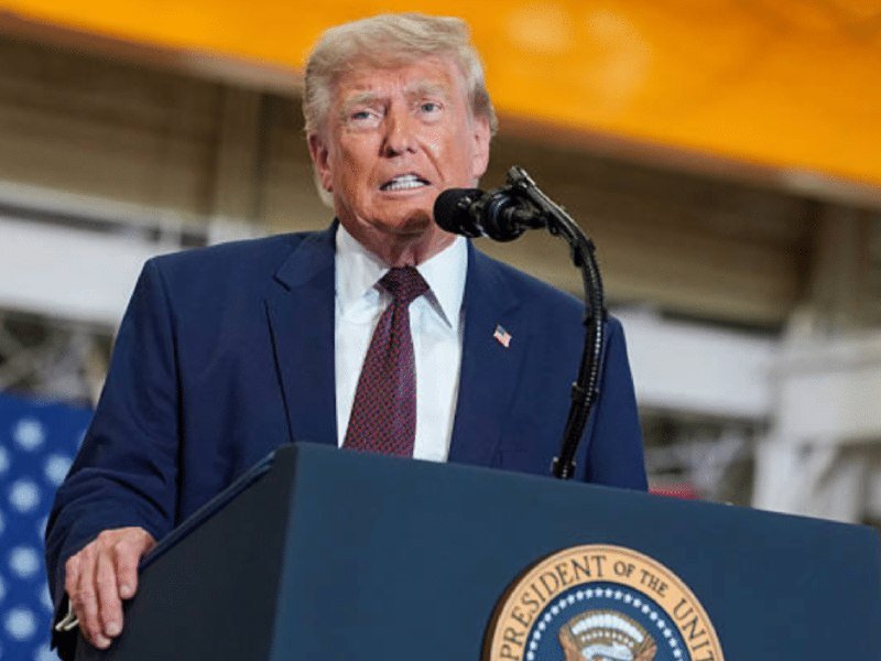 A man in a blue suit and red tie speaks at a podium bearing the presidential seal, with an American flag visible behind him.