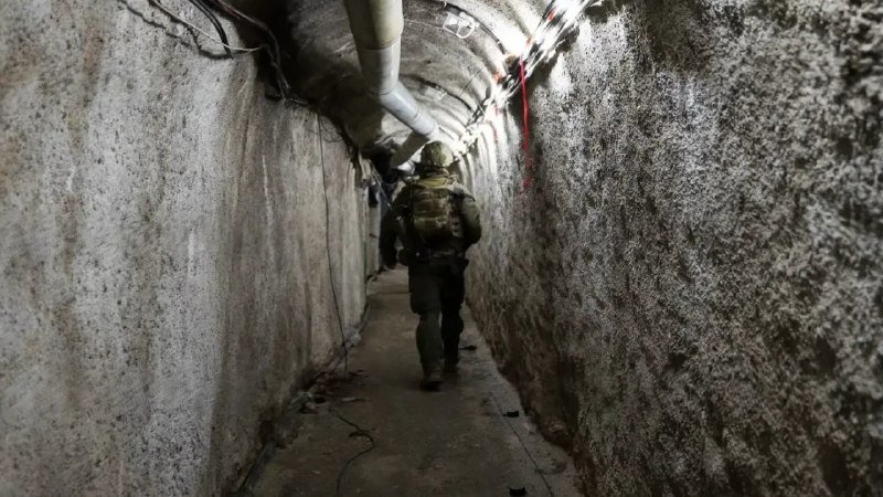 Soldiers in helmets and tactical gear walk single-file through a narrow, dimly lit concrete tunnel with exposed pipes and wiring overhead.