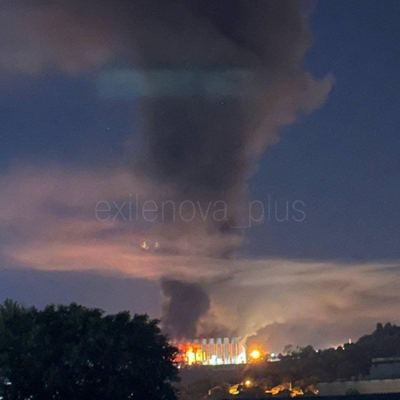 A large plume of dark smoke rises into a twilight sky above a building engulfed in visible flames, with trees silhouetted in the foreground.