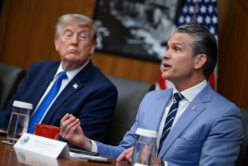 Two men in business suits sit at a conference table with water bottles and name placards, an American flag visible behind them.