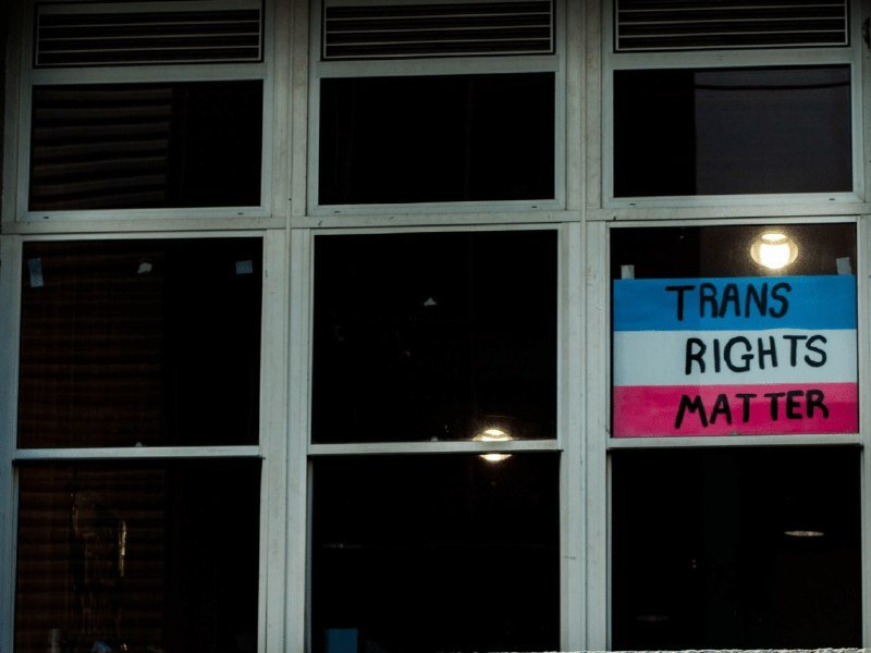 A sign reading "TRANS RIGHTS MATTER" in blue, white, and pink stripes is displayed on the window pane of a building.