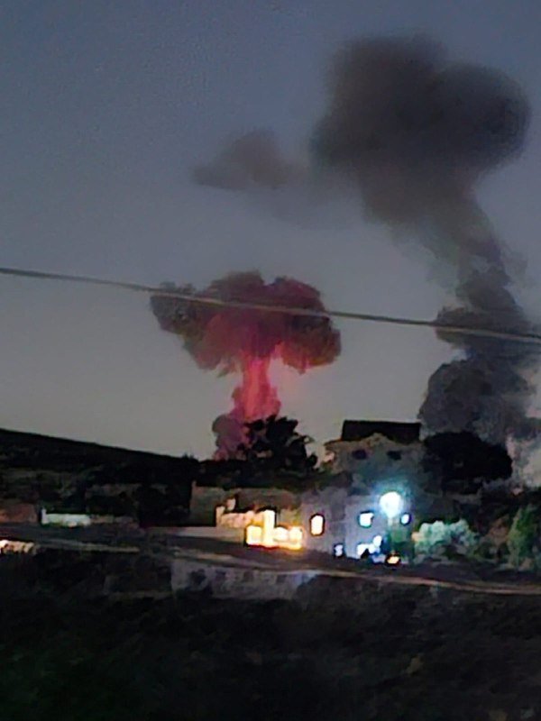 A large pink-tinged mushroom-shaped cloud and a dark plume of smoke rise above buildings at dusk, with power lines crossing the sky and lights glowing below.