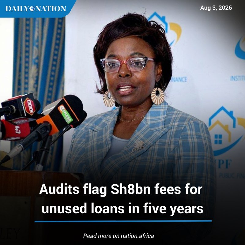 A woman wearing glasses, a blue plaid blazer, and fan-shaped earrings speaks at a podium with multiple microphones, with a "PF PUBLIC FINANCE" logo backdrop and headline about Sh8bn audit fees.
