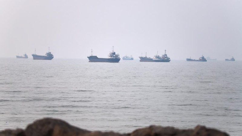 A line of cargo ships sits anchored on a calm, hazy sea, with dark rocks visible in the foreground.