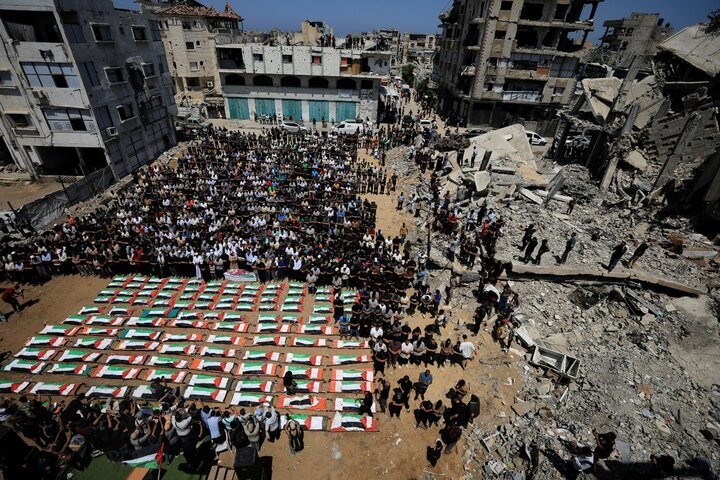 An aerial view shows a large crowd gathered in rows beside dozens of Palestinian flag-draped coffins, with heavily damaged and collapsed buildings visible on the right.