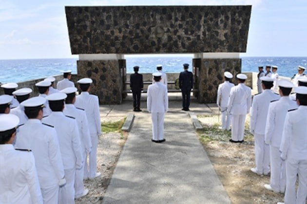 Naval personnel in white uniforms stand in formation facing a large stone monument overlooking the sea during a ceremony.