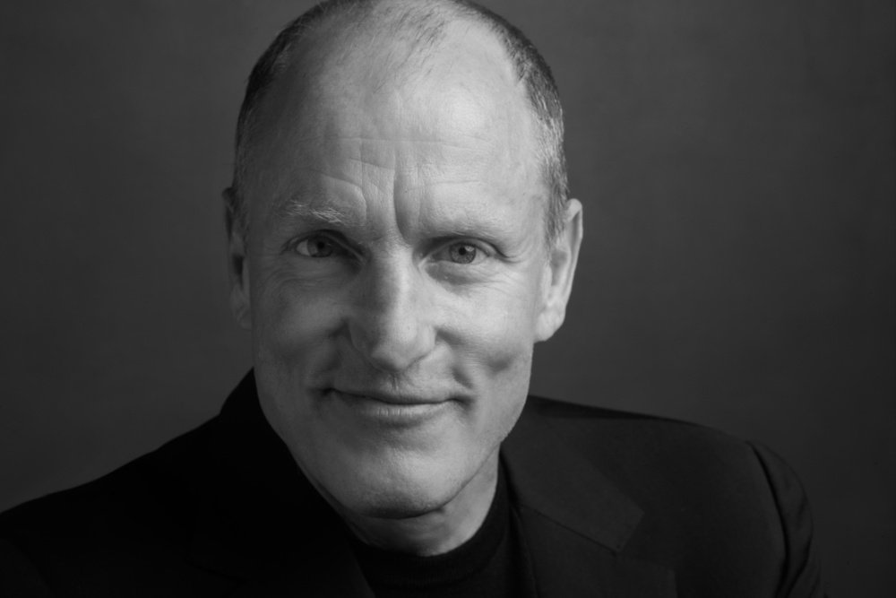 A black-and-white portrait shows a smiling, balding man in a dark jacket posing against a gray background.