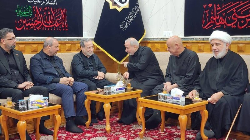 Six men sit conversing around wooden tables in a room decorated with black banners featuring Arabic calligraphy and a central emblem.