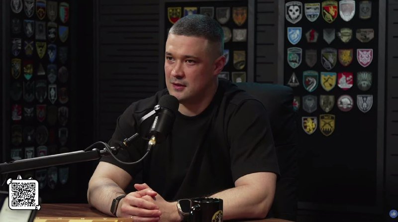 A man in a black t-shirt sits at a wooden table speaking into a microphone, with walls covered in military patches displayed behind him.