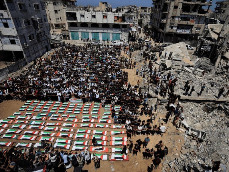 A large crowd gathers around rows of bodies wrapped in Palestinian flags laid out on rubble-strewn ground amid heavily damaged buildings.