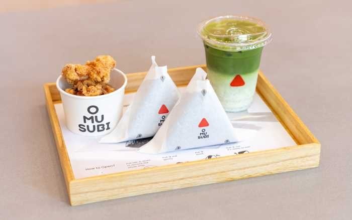 A wooden tray holds a cup of fried chicken, two triangular wrapped packages, and a layered green drink in a clear cup, all branded with the "OMUSUBI" logo.