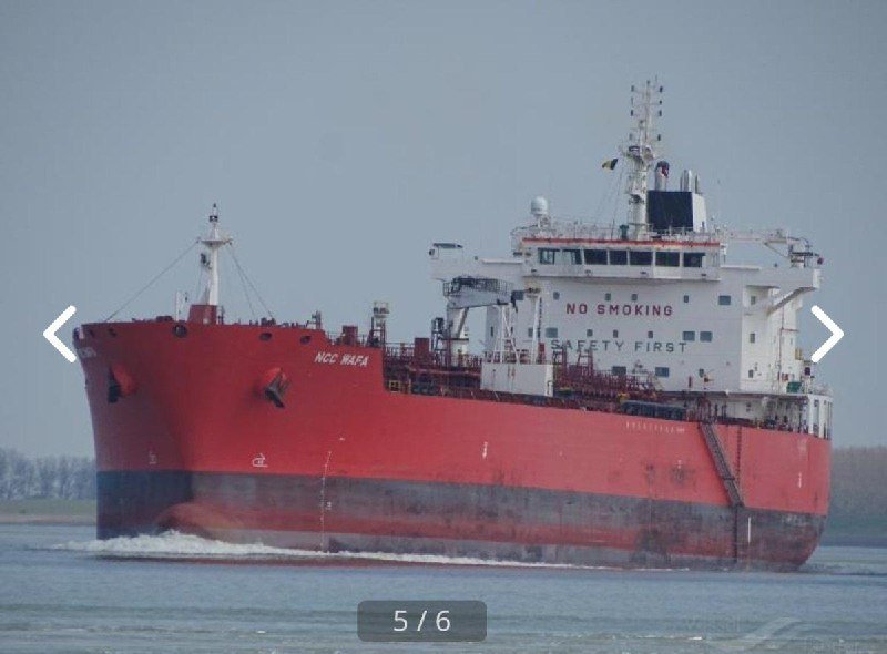 A large red and white cargo ship labeled "NCC WAFA" sails through calm waters, with "NO SMOKING" and "SAFETY FIRST" visible on its white superstructure.