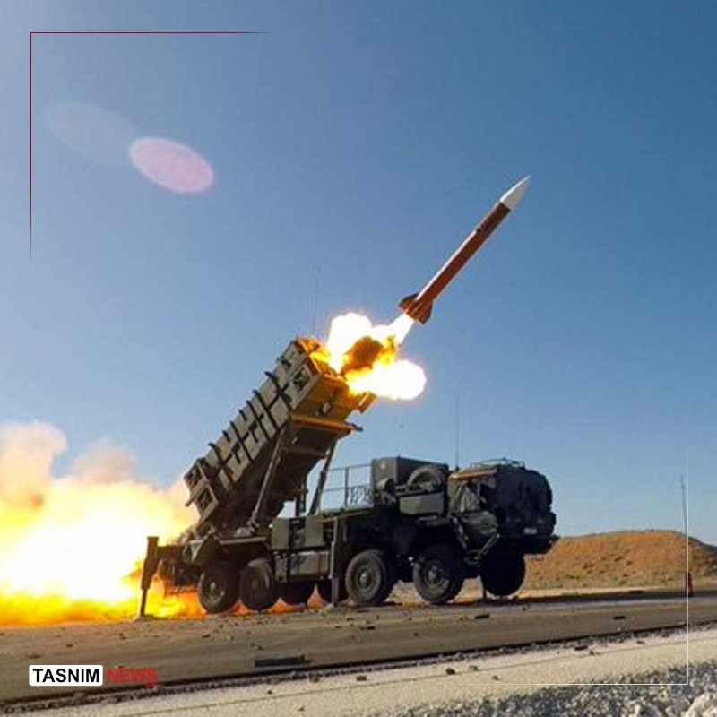 A military truck-mounted launcher fires a missile into a clear blue sky, with bright flames erupting from the launch tube and ground below.