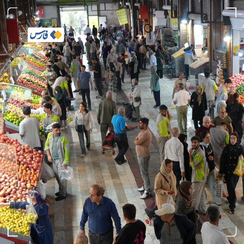 A crowded indoor market shows numerous people walking along a tiled aisle lined with fruit and vegetable stalls.