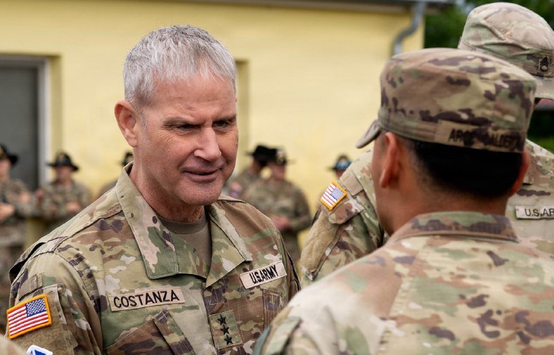 A gray-haired U.S. Army officer with a "COSTANZA" name tape and two stars smiles while speaking with a soldier in a camouflage cap, as other uniformed personnel stand in the background.