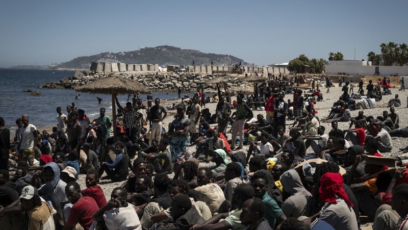 A large crowd of people sits and stands on a rocky beach beside the sea, with a hillside, buildings, and thatched shelters visible in the background under a clear sky.