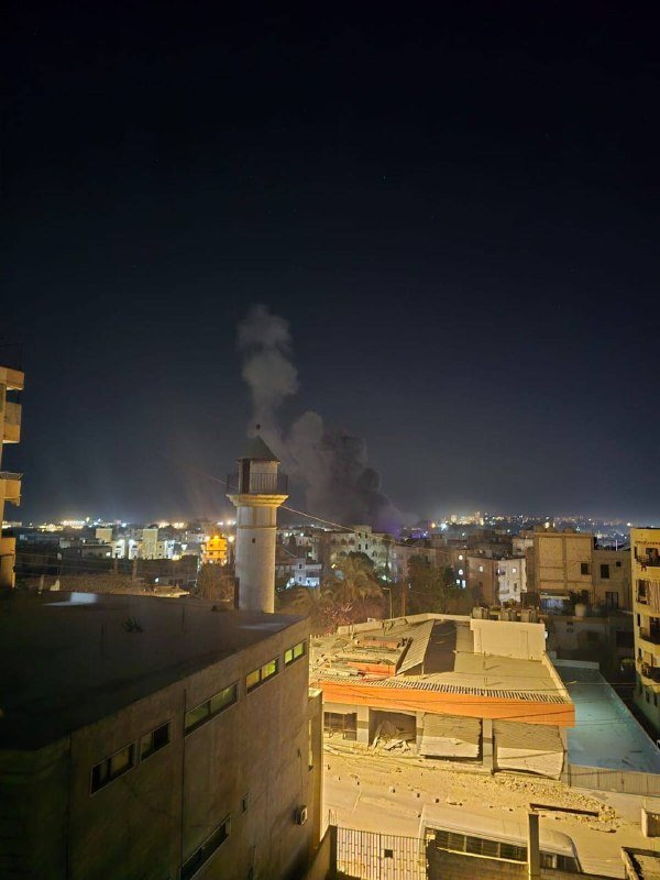 Plumes of smoke rise over a nighttime cityscape featuring a minaret and damaged buildings, with debris scattered across rooftops in the foreground.