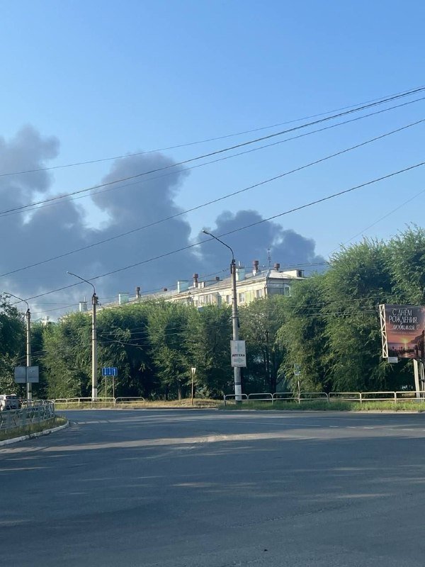 Dark smoke rises behind a residential building and trees along an empty road with overhead power lines.